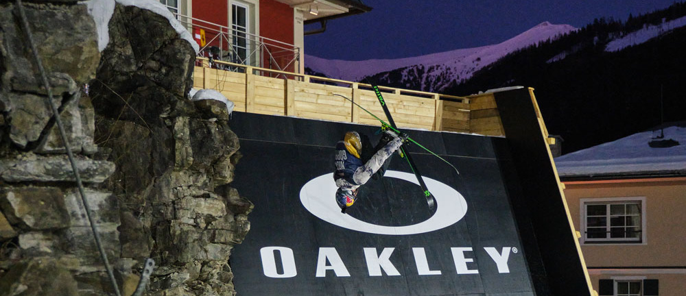 Skifahrer macht einen Trick an einer Rampe mit Häusern und Bergen im Hintergrund