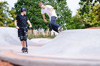 Kind auf Scooter schaut Skateboarder im Ortbeton Skatepark zu