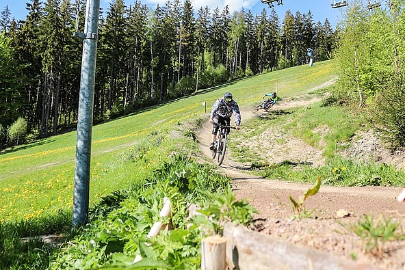 Two bikers at the edge of the forest with chairlift in the background