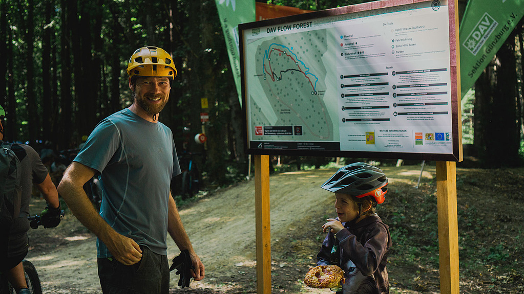 Mann und Kind mit Fahrradhelm stehen vor Mountainbike Infotafel 