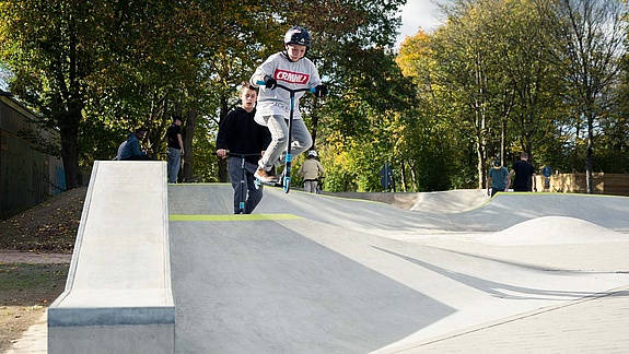 [Translate to Französisch:] Child with scooter jumps in concrete skate park