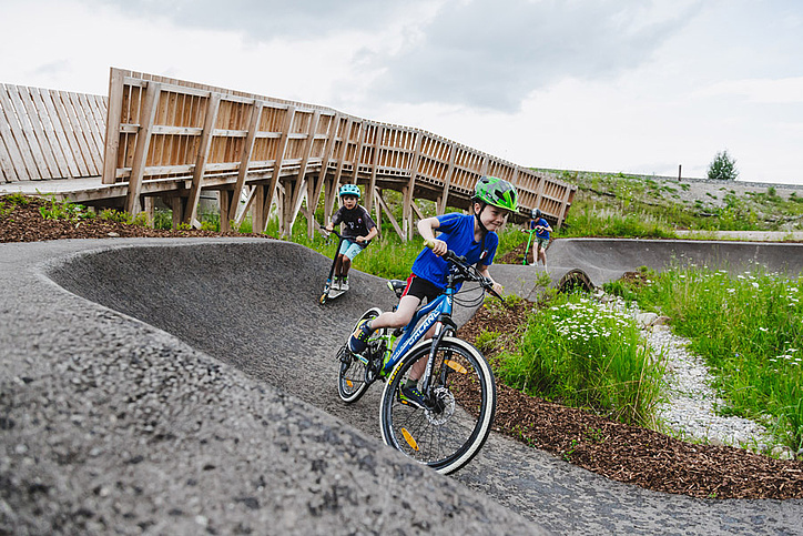 Zwei Kinder fahren mit Bike und Scooter in einer Kurve im Pumptrack