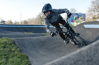 A BMX rider wearing a full-face helmet leans sharply into a paved berm on a pumptrack.