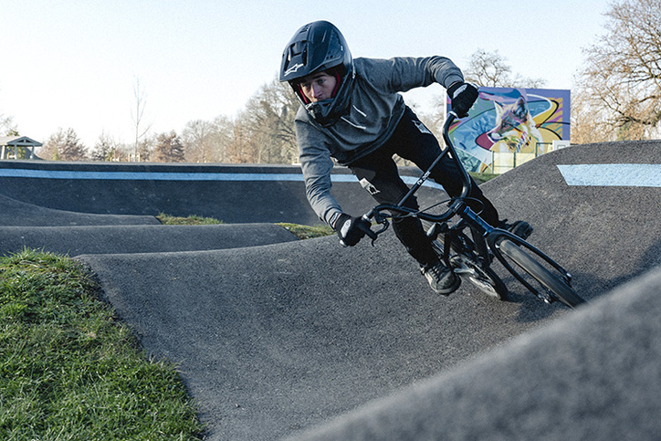 A BMX rider wearing a full-face helmet leans sharply into a paved berm on a pumptrack.