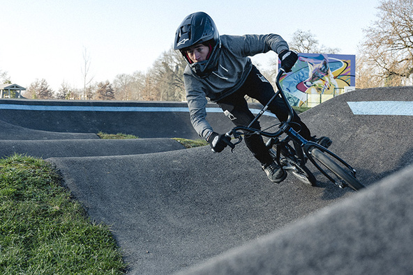 A BMX rider wearing a full-face helmet leans sharply into a paved berm on a pumptrack.