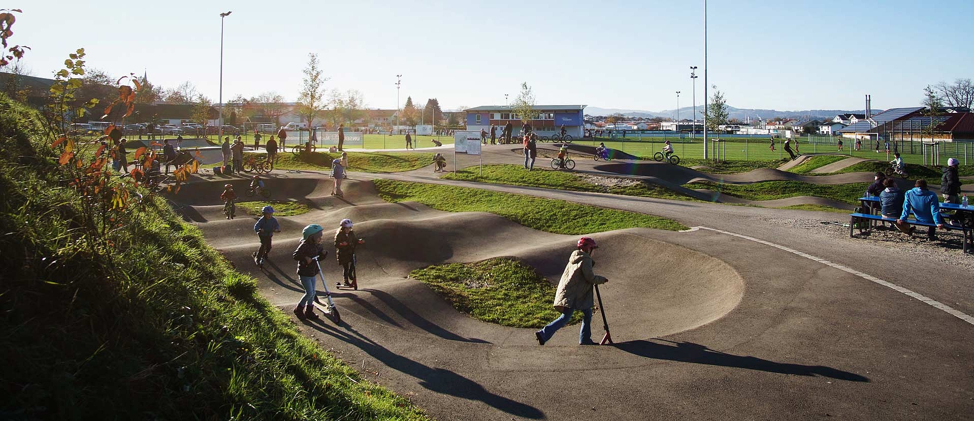Busy pumptrack with kids, scooters and bikes at Urban Sports Park Durach on a sunny day.