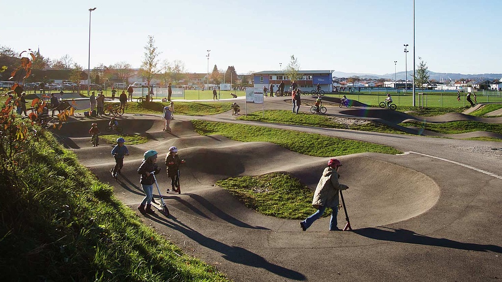 Belebter Pumptrack mit Kindern, Scootern und Fahrrädern im Urban Sports Park Durach bei Sonnenschein.