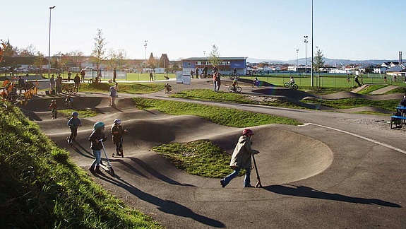 Belebter Pumptrack mit Kindern, Scootern und Fahrrädern im Urban Sports Park Durach bei Sonnenschein.