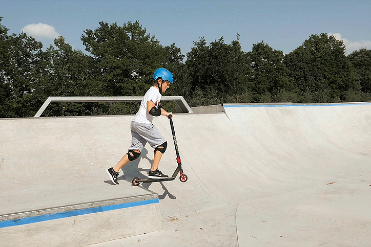 [Translate to Französisch:] Child with scooter drives over heel in skate park