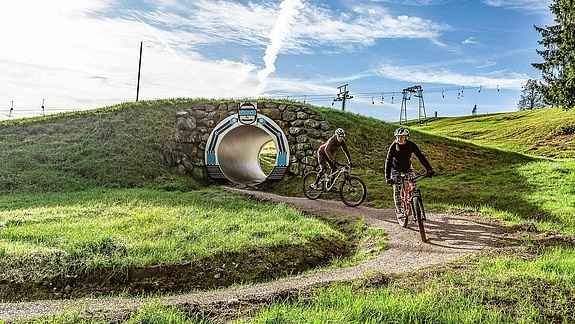 Two female mountain bikers come out of tunnel and ride into curve with lift in background