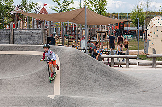 Junge fährt mit Scooter auf Pumptrack im Hintergrund sind Menschen am Tisch