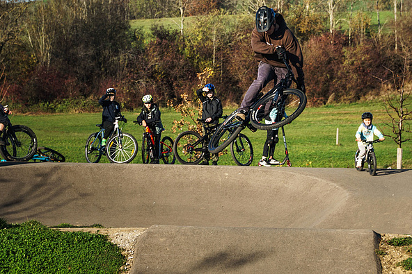 Teen jumps with a bike over a pumptrack wave, spectators in the background at Urban Sports Park Durach.