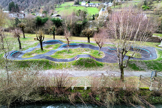 Luftaufnahme des Pumptracks in Saint-Béat-Lez am Flussufer, im Hintergrund entlaubte Bäume und Berglandschaft.