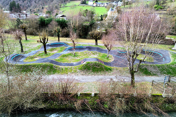 Luftaufnahme des Pumptracks in Saint-Béat-Lez am Flussufer, im Hintergrund entlaubte Bäume und Berglandschaft.