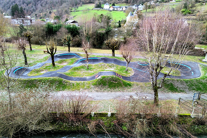Luftaufnahme des Pumptracks in Saint-Béat-Lez am Flussufer, im Hintergrund entlaubte Bäume und Berglandschaft.