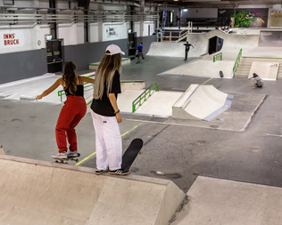 Zwei Skaterinnen in der Skatehalle Innsbruck, umgeben von hochwertigen Holz- und Betonrampen.
