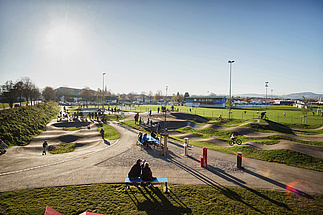 Expansive pumptrack at Urban Sports Park Durach with kids, parents and cyclists in the late afternoon sun.