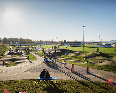 Weitläufiger Pumptrack im Urban Sports Park Durach mit Kindern, Eltern und Fahrradfahrern bei tief stehender Sonne.