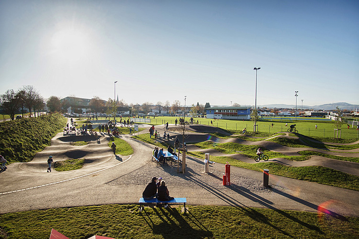 Expansive pumptrack at Urban Sports Park Durach with kids, parents and cyclists in the late afternoon sun.