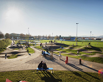 Weitläufiger Pumptrack im Urban Sports Park Durach mit Kindern, Eltern und Fahrradfahrern bei tief stehender Sonne.