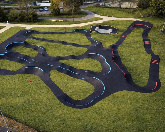 Installation de Pumptrack à Lourdes Grande piste de pumptrack en enrobé à Lourdes avec virages et bosses, entourée d'une végétation dense et d'arbres.