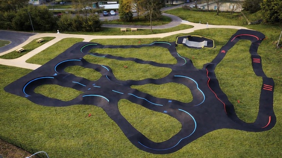 Große, moderne Pumptrack-Anlage in Lourdes mit asphaltierten Kurven und Wellen vor einer Kulisse aus Bäumen und bewaldetem Gelände.
