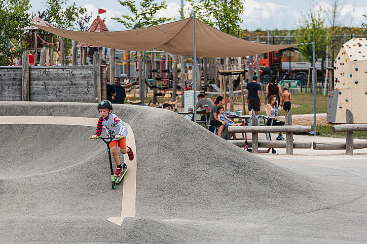 Boy riding scooter on pump track in background are people at table