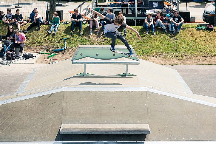 Skateboarder mit blau-weißem T-Shirt springt über eine Box in einem Skatepark