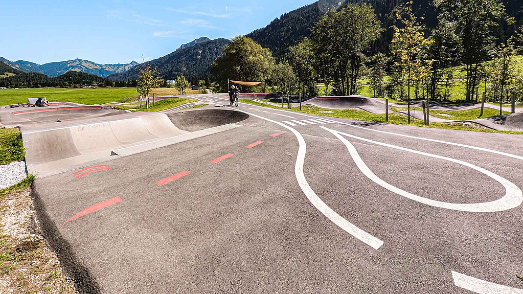 Wide view of the Grän pump track: asphalt rollers with white road markings and a red line, riders on the track, green meadows and a mountain range under a clear blue sky.