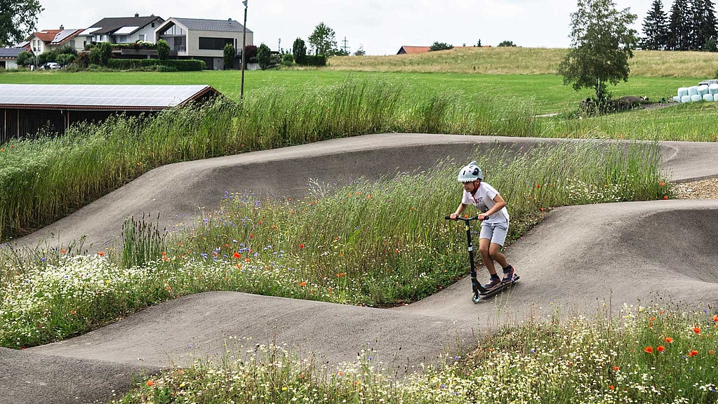  Junge mit Scooter fährt durch blumenreichen, naturnah gestalteten Pumptrack im Urban Sports Park Durach.