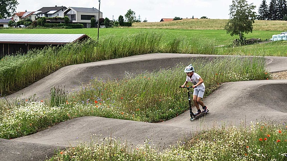 Boy on a scooter rides through a flower-rich, nature-integrated pumptrack at Urban Sports Park Durach.