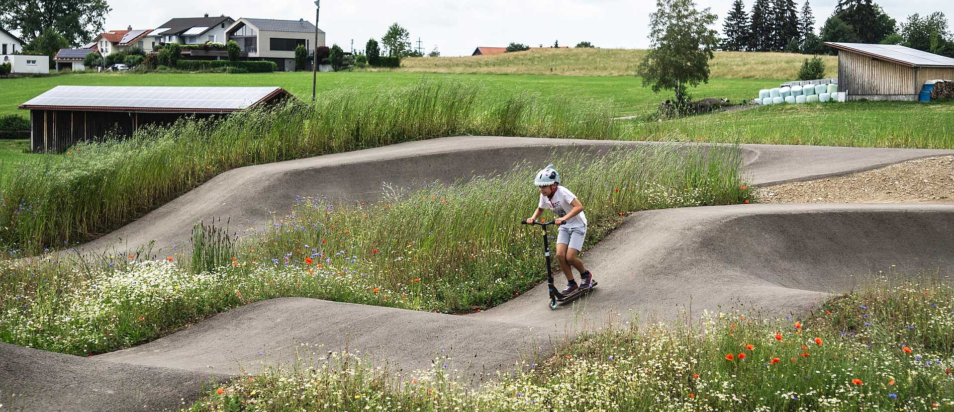 Boy on a scooter rides through a flower-rich, nature-integrated pumptrack at Urban Sports Park Durach.