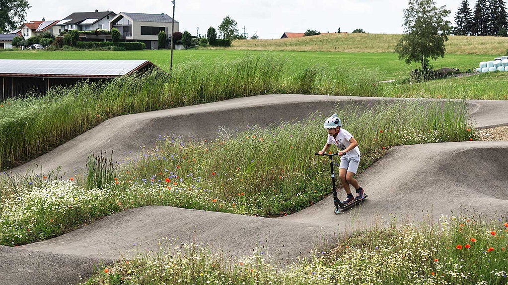 Boy on a scooter rides through a flower-rich, nature-integrated pumptrack at Urban Sports Park Durach.