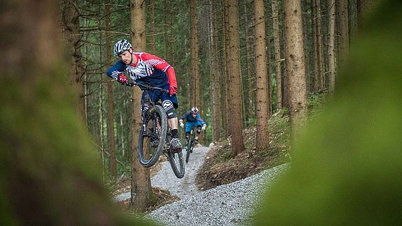 Two bikers jump one after the other on prepared trail in forest