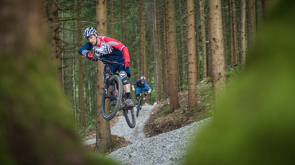 Two bikers jump one after the other on prepared trail in forest