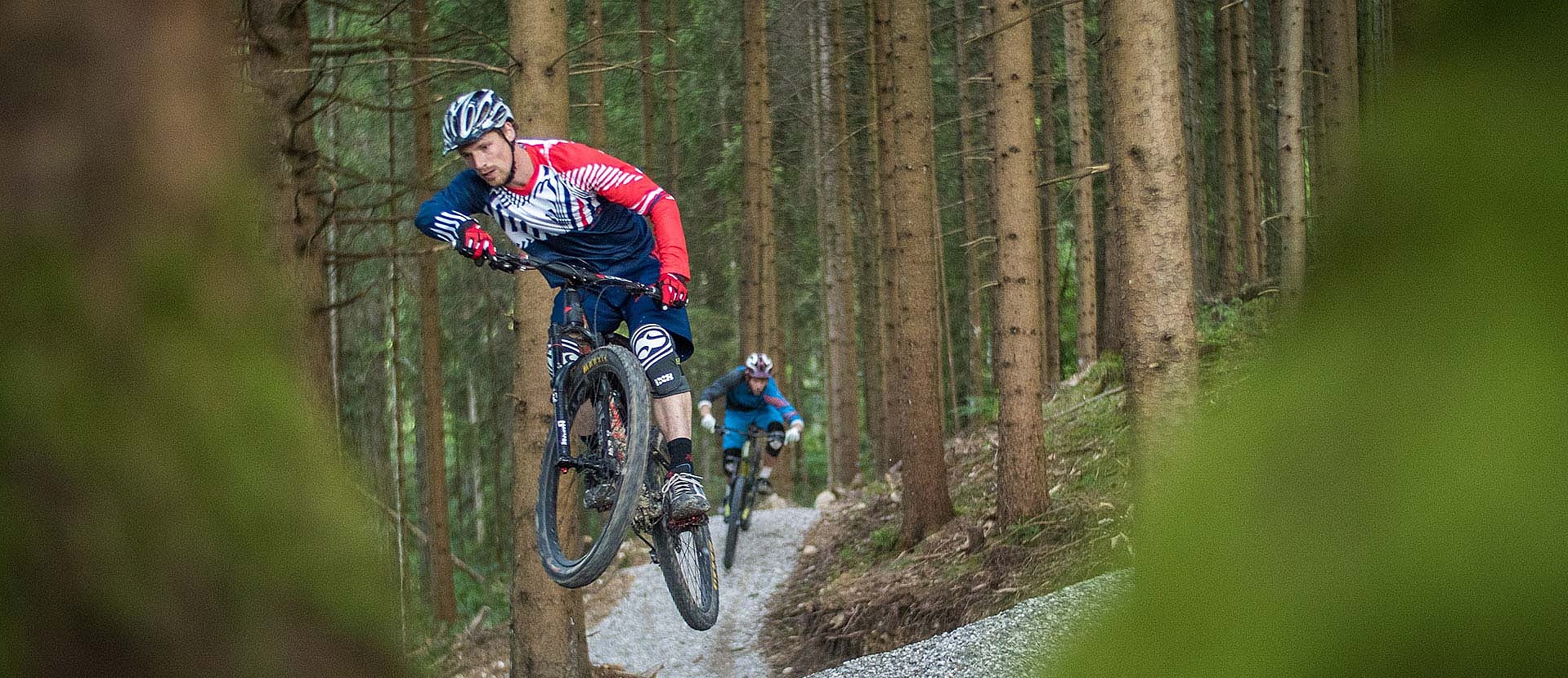Two bikers jump one after the other on prepared trail in forest