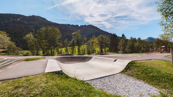 Scooterloop mit Ein- und Ausfahrt neben der Asphaltfläche des Pumptracks; davor Gras und Schotter, dahinter Wald und Berge unter leicht bewölktem Himmel.