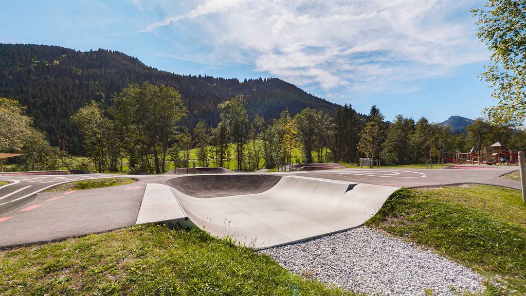Scooterloop mit Ein- und Ausfahrt neben der Asphaltfläche des Pumptracks; davor Gras und Schotter, dahinter Wald und Berge unter leicht bewölktem Himmel.