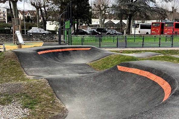 Berm of a pumptrack in Saint-Girons with orange lines, next to a playground and parked buses.