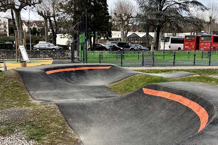 Berm of a pumptrack in Saint-Girons with orange lines, next to a playground and parked buses.
