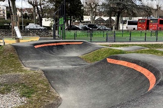 Berm of a pumptrack in Saint-Girons with orange lines, next to a playground and parked buses.