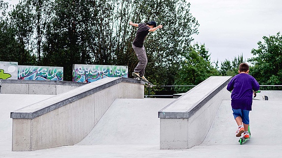 Child with scooter watches skateboarder do a trick in a in-situ concrete skatepark