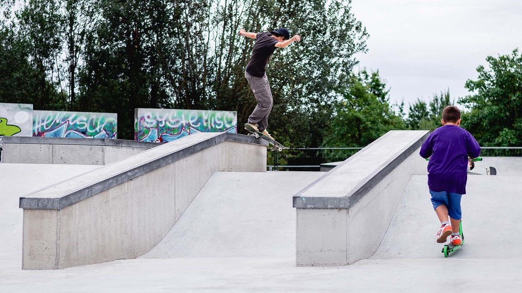 Kind mit Scooter schaut Skateboarder bei einem Trick in einem Ortbeton Skatepark zu