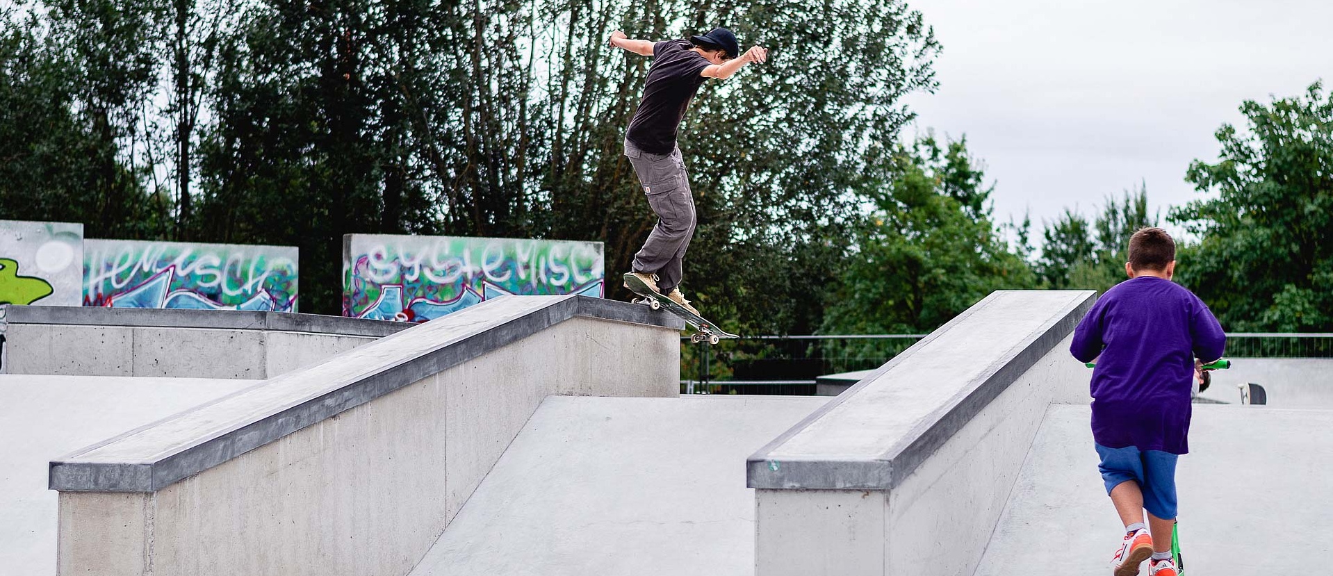 Kind mit Scooter schaut Skateboarder bei einem Trick in einem Ortbeton Skatepark zu