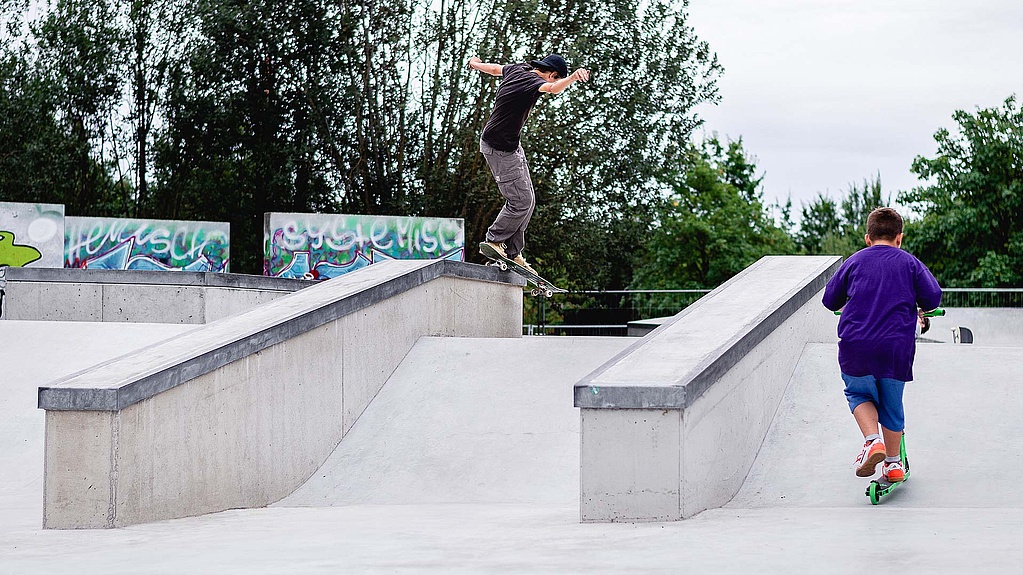 Child with scooter watches skateboarder do a trick in a in-situ concrete skatepark
