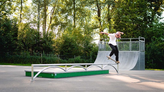 Skateboarder performing a trick on a flat rail at Isny ramp park with a forest background.