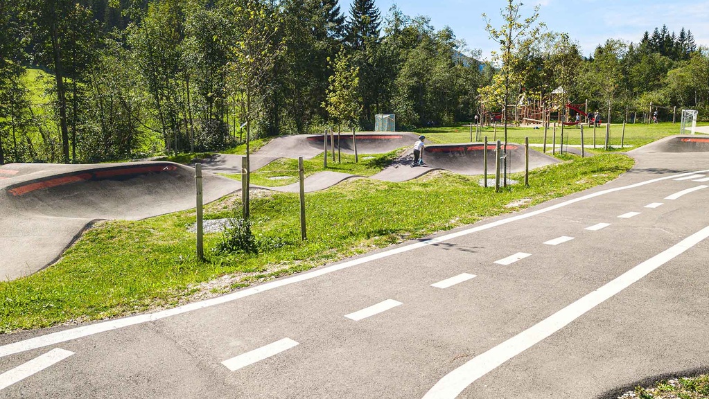 Abschnitt des Pumptracks mit niedrigen Wellen und Steilkurven, junge Fahrende auf der Strecke; daneben ein Lernverkehrsweg mit weißen Markierungen und im Hintergrund ein Spielplatz.