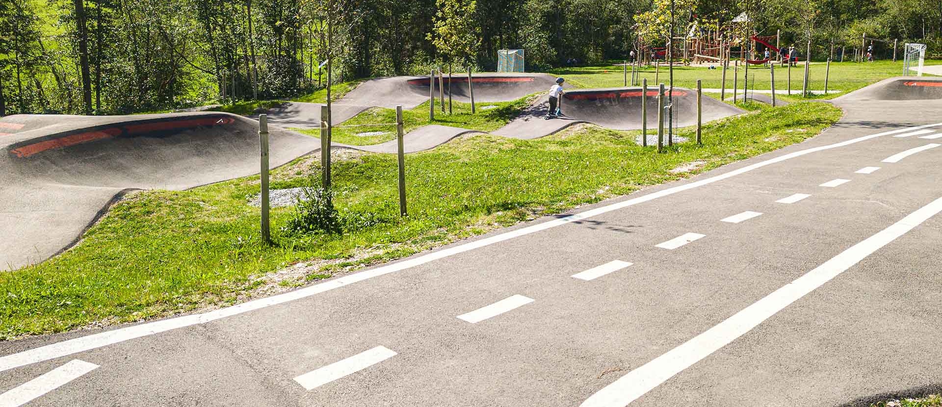 Abschnitt des Pumptracks mit niedrigen Wellen und Steilkurven, junge Fahrende auf der Strecke; daneben ein Lernverkehrsweg mit weißen Markierungen und im Hintergrund ein Spielplatz.