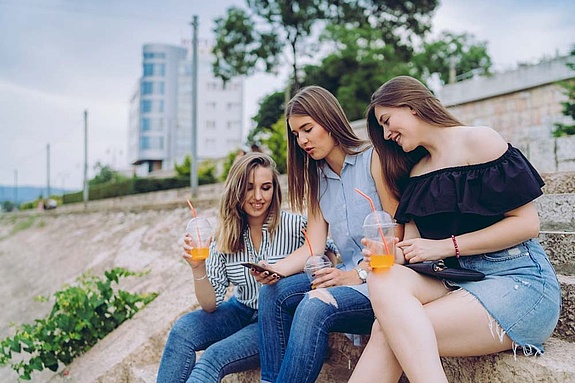 Drei Mädchen sitzen mit Getränken auf einer Steintreppe in urbaner Umgebung