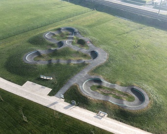 [Translate to Französisch:] Luftaufnahme eines modernen, geschwungenen Asphalt-Pumptracks auf einer grünen Wiese neben Bahngleisen.